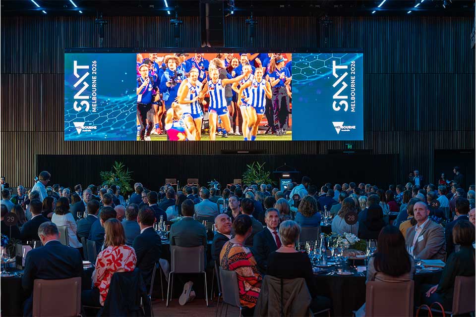 Attendees of the Sport NXT conference sit around tables. A big screen on the wall promotes the Melbourne 2026 event with a picture of the North Melbourne WAFL team celebrating.