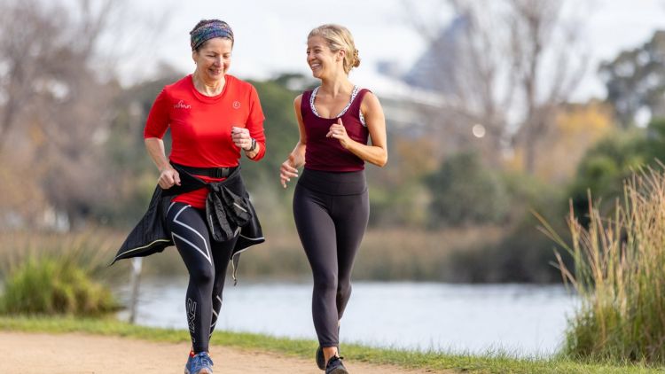 Two smiling women in workout gear run on a gravel path by a lake.