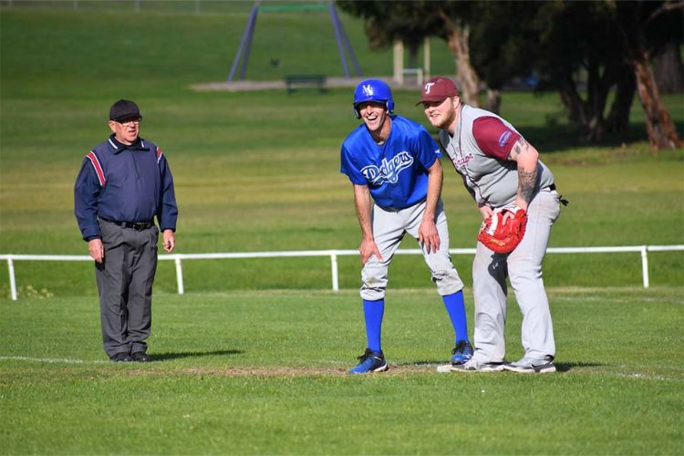 A baseman and runner stand at a base watching the action while an umpire stands in the background.