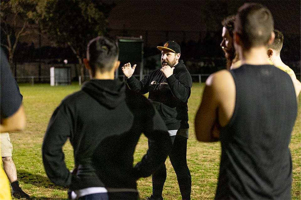 A coach in a black top and cap addresses players at an evening training session.