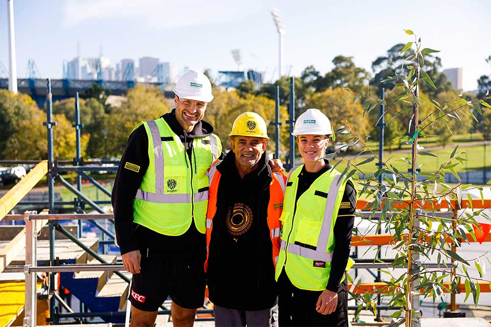 Three people in helmets and high-visibility vest stand arm-in-arm at a construction site.