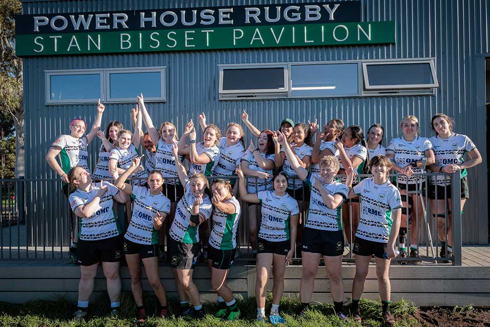 The Power House women's rugby team in uniform, pointing up at a sign reading, 'Power House Rugby Stan Bisset Pavillion'.