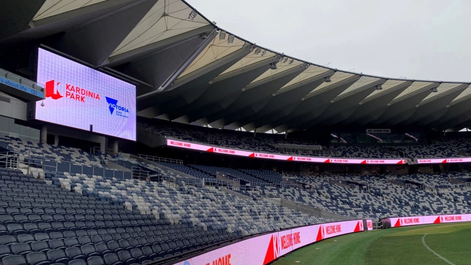 Photo of the Kardinia Park stadium taken from the grass. The seats are empty and a jumbo screen shows the Kardinia Park and Victoria State Government logos.