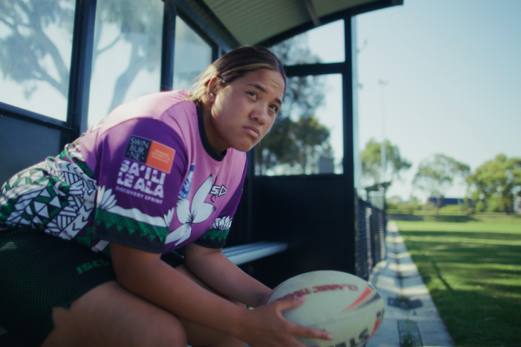 Chloe siting on covered bench next to rugby pitch holding a rugby ball