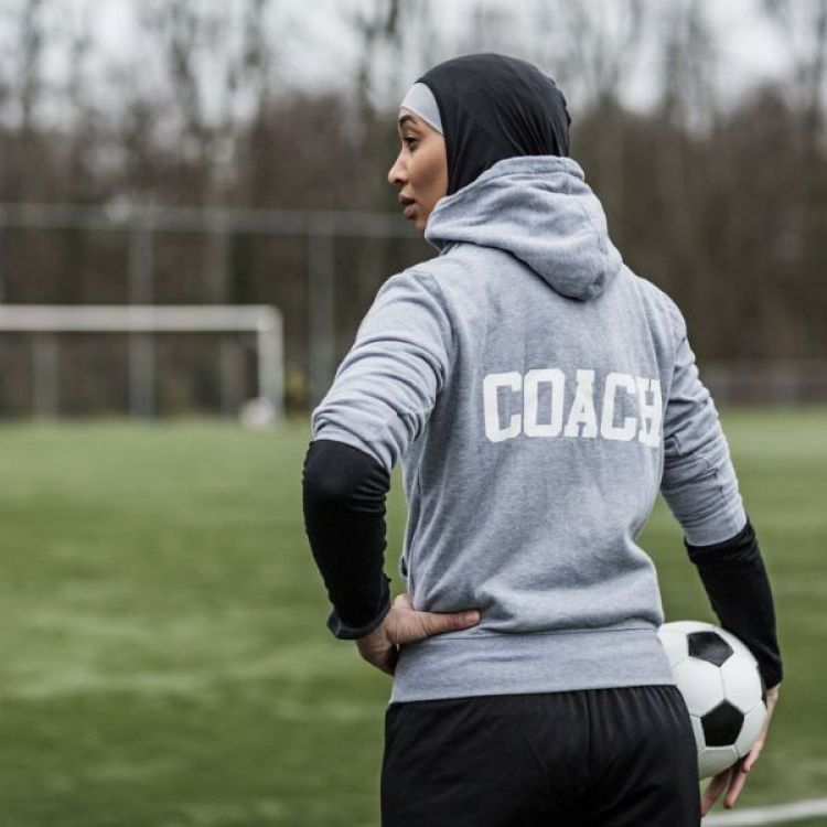 A muslim woman wears a sport hijab and a hoodie with the word 'Coach' on the back.
