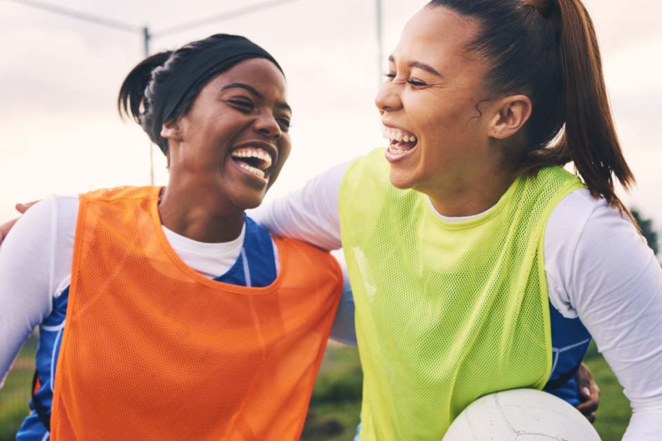 Two women in different coloured vests laugh together. One is holding a soccer ball.