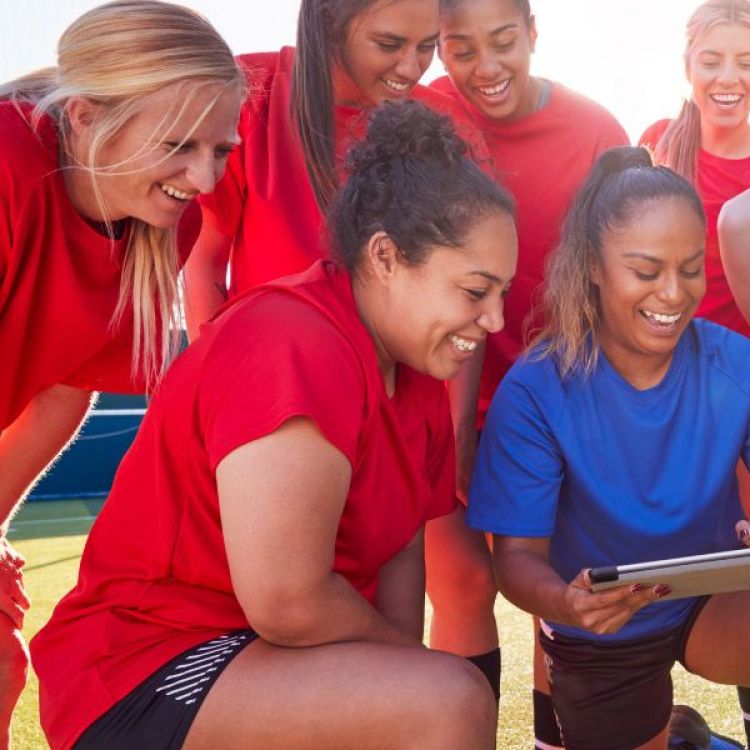 Sportswomen laugh with their coach while looking at something on a tablet.