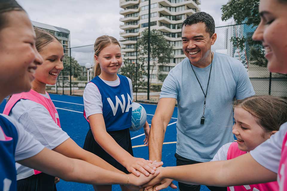 A coach and young netball players wearing bibs showing the letters of their court positions stack their hands together before a game.