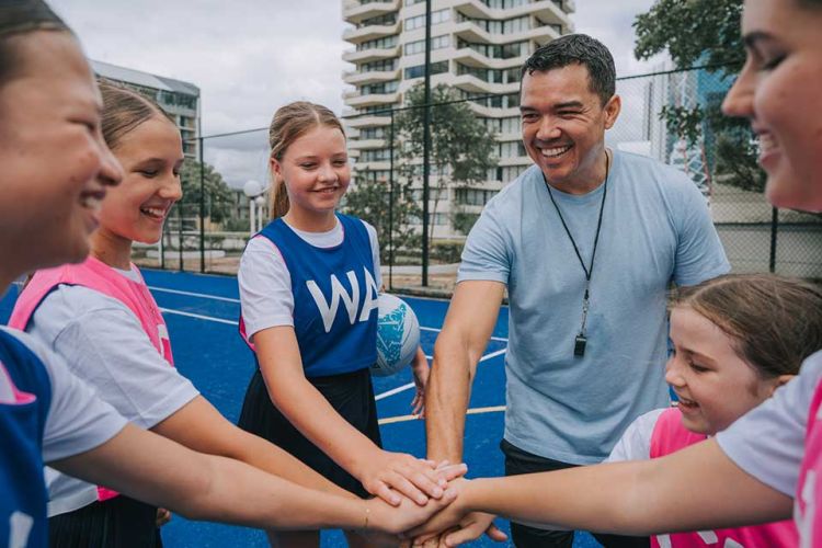 A coach and young netball players wearing bibs showing the letters of their court positions stack their hands together before a game.