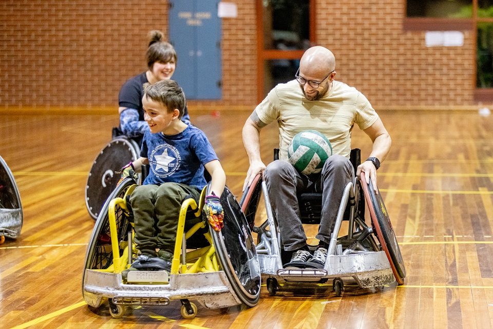 Three smiling people in wheelchairs play a game of rugby indoors.