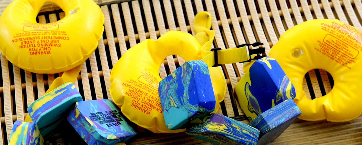 An assortment of floaties and floating belts lie at the edge of a public indoor swimming pool.