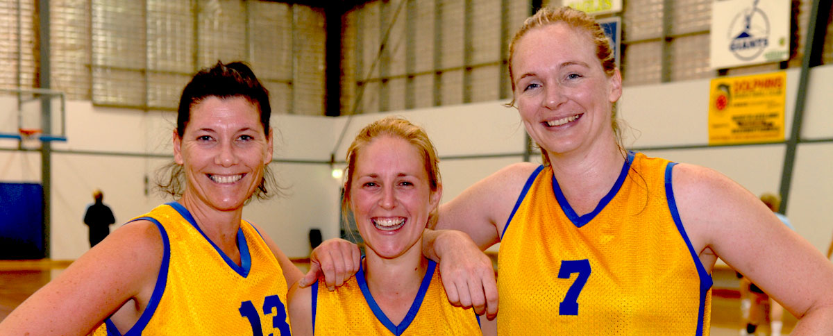 Three female basketballers stand on an indoor basketball court, smiling.